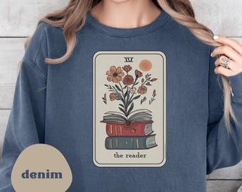 Puede incluir: Sudadera azul denim con un diseño de carta del tarot. La carta muestra una pila de libros, flores y el texto "the reader". La carta tiene un fondo beige con un sol y el número romano IV. La palabra "denim" está impresa en un óvalo beige.
