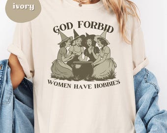 Puede incluir: Camiseta color marfil con un diseño en sepia de cuatro brujas alrededor de un caldero. El texto "GOD FORBID WOMEN HAVE HOBBIES" está impreso. La camiseta tiene un corte holgado y está hecha de un material suave.