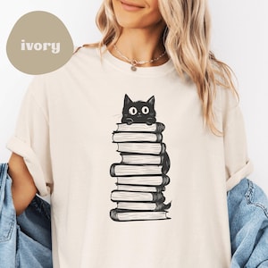 May include: A white t-shirt with a black cat peeking out from behind a stack of books. The cat has large eyes and a surprised expression. The shirt is a unisex style and is available in a variety of sizes.