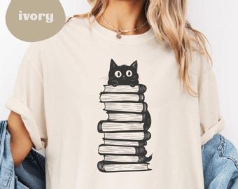 Könnte beinhalten: Ein weißes T-Shirt mit einer schwarzen Katze, die aus einem Stapel Bücher schaut. Die Katze hat große Augen und einen überraschten Ausdruck. Das Shirt ist unisex und in verschiedenen Größen erhältlich.