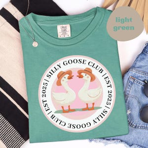 Puede incluir: Camiseta verde claro con un diseño circular de dos gansos con sombreros y el texto "SILLY GOOSE CLUB | EST 2025". La camiseta está hecha de un material suave y es cómoda.