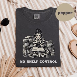 May include: Dark grey t-shirt with a graphic of a woman surrounded by books, reading a book. The text "NO SHELF CONTROL" is printed below the graphic. The shirt is a Comfort Colors brand.