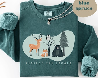 May include: A blue spruce sweatshirt with a nature-themed graphic. The graphic features a deer, fox, owl, and bear, with a tree and the text "RESPECT THE LOCALS". A silver necklace with a pendant is draped over the sweatshirt.