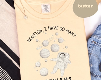 May include: A light yellow t-shirt with a black and white illustration of a cat in a spacesuit surrounded by planets. The text "HOUSTON, I HAVE SO MANY PROBLEMS" is printed on the shirt.