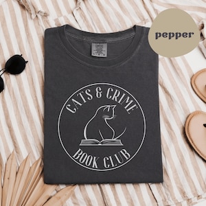 May include: A dark gray t-shirt with the text "CATS & CRIME BOOK CLUB" in a white circular design. The design includes a cat silhouette sitting on an open book. The shirt is laid flat on a striped surface.