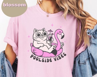 Könnte beinhalten: Hellrosa T-Shirt mit einer Grafik einer Katze mit Sonnenbrille in Blumenform, die auf einem rosa Flamingo-Schwimmer mit einem Cocktail liegt. Der Text "Poolside Vibes" steht darunter. Das Wort "blossom" befindet sich in einem hellbraunen Kreis.