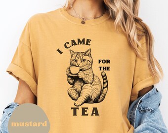 Op de afbeelding: Mosterdkleurig T-shirt met een afbeelding van een kat die een theekopje vasthoudt, met de tekst "I CAME FOR THE TEA". Het shirt heeft opgerolde mouwen en een relaxte pasvorm. Het woord "mustard" is gedrukt op een ovale tan.