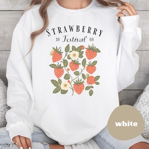 May include: White sweatshirt featuring a strawberry design with the text "STRAWBERRY Festival 69". The design includes red strawberries, green leaves, and white flowers. The sweatshirt is a long-sleeved crew neck.