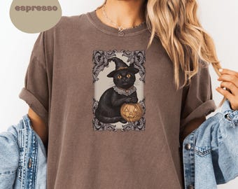 May include: Espresso-colored t-shirt featuring a framed illustration of a black cat wearing a witch hat and holding a carved pumpkin. The cat has a ruffled collar. The shirt has rolled-up sleeves.