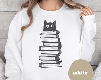 Op de afbeelding: Witte trui met een zwarte kat die uit een stapel boeken kijkt. De kat kijkt naar links. De tekst "white" is gedrukt op een bruine cirkel in de rechterbenedenhoek van de afbeelding.