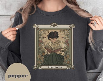 Puede incluir: Sudadera gris oscuro con una ilustración vintage de una mujer leyendo un libro, enmarcada por elementos florales. La ilustración está etiquetada como "the reader". La sudadera tiene un corte relajado y un color neutro.