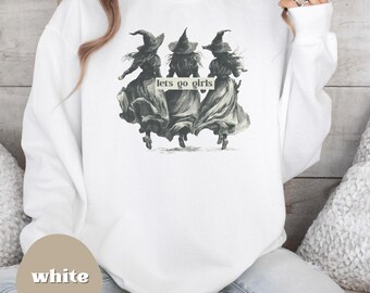May include: White sweatshirt featuring a grayscale illustration of three witches in hats and flowing dresses. The text "lets go girls" is printed on the design. The sweatshirt is a casual, comfortable style.