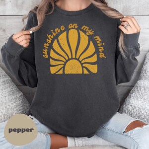 May include: Dark gray sweatshirt with the text "sunshine on my mind" in yellow above a yellow sunburst design. The sweatshirt is a relaxed fit and has long sleeves. The word "pepper" is in a circle at the bottom.