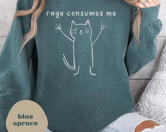 Op de afbeelding: Een tealblauwe trui met de tekst "rage consumes me" en een witte lijntekening van een kat met zijn armen omhoog. De trui is gemaakt van een zacht, comfortabel materiaal.