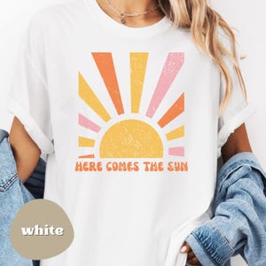 May include: White t-shirt featuring a retro sun graphic in shades of orange, pink, and yellow. The phrase "HERE COMES THE SUN" is printed in orange below the sun design. The shirt has rolled-up sleeves.