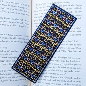 May include: A rectangular fabric bookmark featuring a repeating pattern of embroidered mushrooms in yellow, pink, and blue thread on a black background, placed diagonally across the open pages of a printed book with visible text.
