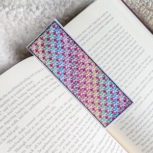 May include: A cross-stitched bookmark featuring a floral pattern in pink, blue, and green. The bookmark is rectangular and sits within the pages of a book, offering a handcrafted touch to reading.