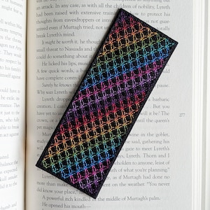 Op de afbeelding: Een rechthoekige bladwijzer met een geometrisch regenboogpatroon, liggend op de open pagina's van een boek. Het ontwerp van de bladwijzer heeft een herhalend ruitpatroon in verschillende kleuren.