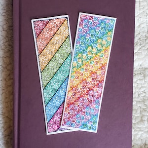 Può includere: Due segnalibri fatti a mano con un motivo arcobaleno, appoggiati su un libro viola. I segnalibri presentano un design diagonale con colori come rosso, arancione, giallo, verde, blu e viola. Il libro ha una finitura liscia e opaca.