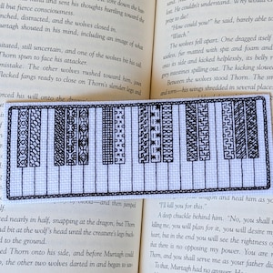 May include: A rectangular white fabric bookmark featuring a black embroidered piano keyboard design. The black keys are decorated with various intricate geometric patterns, while the white keys are outlined in black thread on a plain white background.