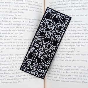 May include: A rectangular black bookmark featuring a white embroidered pattern of various ghost figures and the repeated text "BOO!" throughout the design. The bookmark has a stitched border and is placed against an open book page.
