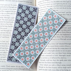 May include: Two rectangular bookmarks with floral patterns. One bookmark has a white background with a pattern of blue and black flowers. The other has a white background with red and blue flowers. Both are on a book page.
