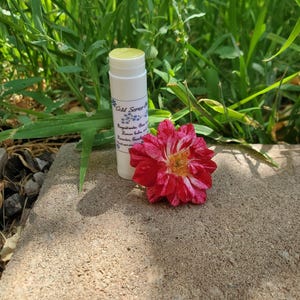May include: A white lip balm tube with the text "Cold Sores" and a red and white flower. The lip balm is next to a red and white flower on a concrete surface, with green grass in the background.
