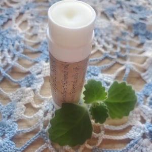 May include: A white lip balm stick with a label featuring text, Sweet Almond oil, and Essential oils. The balm is placed on a blue and white crocheted surface, with green leaves nearby.