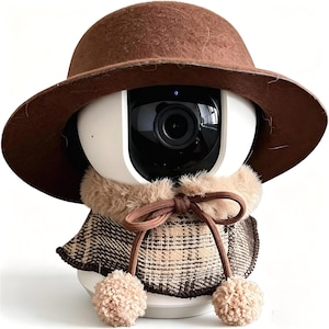 May include: A white spherical security camera wearing a brown felt hat, a beige faux fur collar, and a brown and beige plaid capelet tied with a drawstring featuring two beige pom-poms at the ends of the strings.