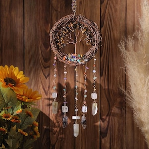 Puede incluir: Un atrapasueños de rama marrón con un diseño de árbol de la vida y acentos de piedras preciosas de colores. El atrapasueños tiene múltiples hilos de cuentas y cristales que cuelgan de él.