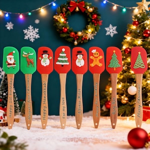 May include: A collection of colorful Christmas-themed spatulas with wooden handles. Each spatula features a different holiday design, including Santa Claus, a reindeer, a snowman, a Christmas tree, and a gingerbread man. The handles have holiday greetings.