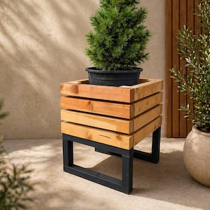 May include: A small evergreen tree in a black pot sits on a wooden planter box with a slatted design. The planter is supported by a black metal frame. The wood has a natural, light brown colour.