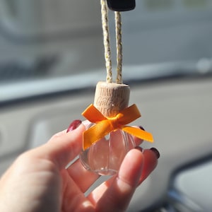 May include: A small, clear glass car air freshener bottle with a light-colored wooden cap engraved with the words Thankful, Grateful, Blessed. The bottle is adorned with a bright orange ribbon and hangs from a gold and white braided cord.