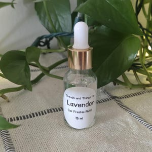 May include: A clear glass bottle with a gold-colored dropper cap containing liquid. The white label reads: Threads and Things Co. Lavender Car Freshie Refill 15 ml. The bottle has a white dropper tip and is filled with a transparent liquid substance.