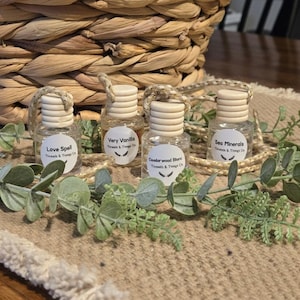 May include: Four small glass bottles with wooden caps and labels reading "Love Spell", "Very Vanilla", "Cedarwood Blend", and "Sea Minerals". The bottles are arranged on a beige surface with green foliage and a woven basket in the background.