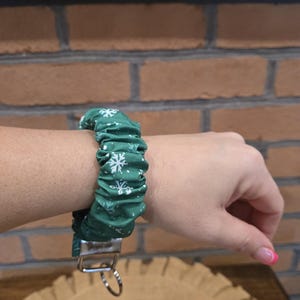 May include: A dark green fabric wristlet keychain featuring a repeating white snowflake pattern. The accessory has a gathered, scrunchie-style design and includes a silver-toned metal hardware clasp with an attached circular key ring.