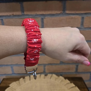 May include: A red fabric wristlet keychain featuring a repeating white snowflake pattern. The gathered fabric is attached to a silver-toned metal hardware clasp with a circular split ring for keys, designed to be worn around the wrist.