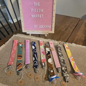May include: A collection of baby pacifier clips with various fabric patterns, including floral, sports, and camouflage designs. The clips are displayed on a table with a sign that reads "THE PILLOW MARKET BY LEEANNE". Each clip has a metal clasp and a small tag.