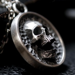 Puede incluir: Un collar con colgante de plata con un diseño detallado de calavera. La calavera está dentro de un marco circular y cuelga de una delicada cadena. El colgante y la cadena son de un material metálico brillante.