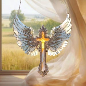 May include: A decorative cross with angel wings, crafted with silver-colored metal and white and yellow stained glass. The cross is suspended in front of a window with a view of a field.