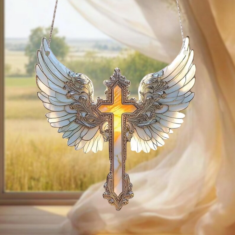May include: A decorative cross with angel wings, crafted with silver-colored metal and white and yellow stained glass. The cross is suspended in front of a window with a view of a field.