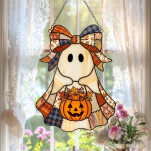 May include: A stained glass suncatcher shaped like a ghost holding a jack-o'-lantern filled with autumn leaves and berries. The ghost features a large bow at the top with a patchwork pattern of orange, blue, and white plaid and floral prints.