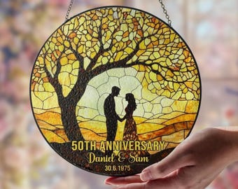 May include: A round stained glass piece with a tree and couple silhouette design. The glass is yellow and orange with a black outline. Text reads "50th Anniversary Daniel & Sam 30.6.1975".