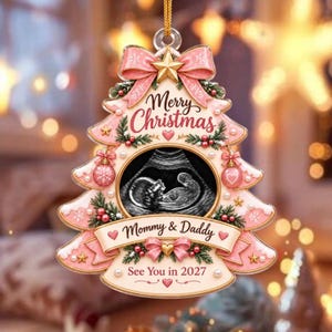 May include: A Christmas tree-shaped ornament in shades of pink and cream, featuring a central ultrasound image, a gold star, pink bows, and text that reads Merry Christmas, Mommy & Daddy, and See You in 2027, suspended by a gold cord.