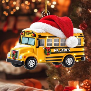 May include: A yellow school bus Christmas ornament featuring a red Santa hat with white trim on the roof, a green wreath on the front grille and side, and the text "SCHOOL BUS" and "MR. JAMES" printed on the side of the vehicle.
