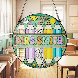 May include: A round, stained-glass window hanging with a black chain. The design features colorful pencils, a paper airplane, a paper clip, and the name "MRS. SMITH" in a retro font. The background is a classroom.