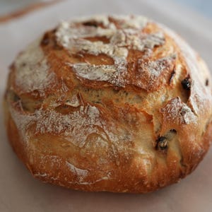 Könnte beinhalten: Ein rundes Brot aus handwerklicher Herstellung mit goldbrauner Kruste, bestäubt mit Mehl. Das Brot hat ein rustikales Aussehen und liegt auf einem weißen Pergamentpapier. Kleine, dunkle Einschlüsse sind sichtbar.