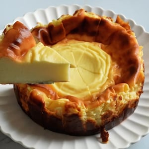 Puede incluir: Un pastel de queso entero con una corteza dorada y un centro amarillo claro, presentado en un plato blanco con borde festoneado. Una porción se levanta con un servidor plateado, mostrando la textura del pastel.