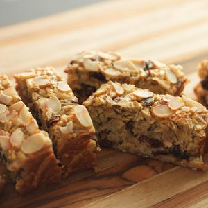 Puede incluir: Primer plano de cuatro rebanadas de una barra de granola con almendras en la parte superior. Las barras de granola están sobre una tabla de cortar de madera.