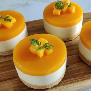 Op de afbeelding: Close-up van verschillende mini-mangocheesecakes op een houten plank. Elke cake heeft een witte basis, een laag feloranje glazuur en is gegarneerd met mangoblokjes en een takje groene garnering.